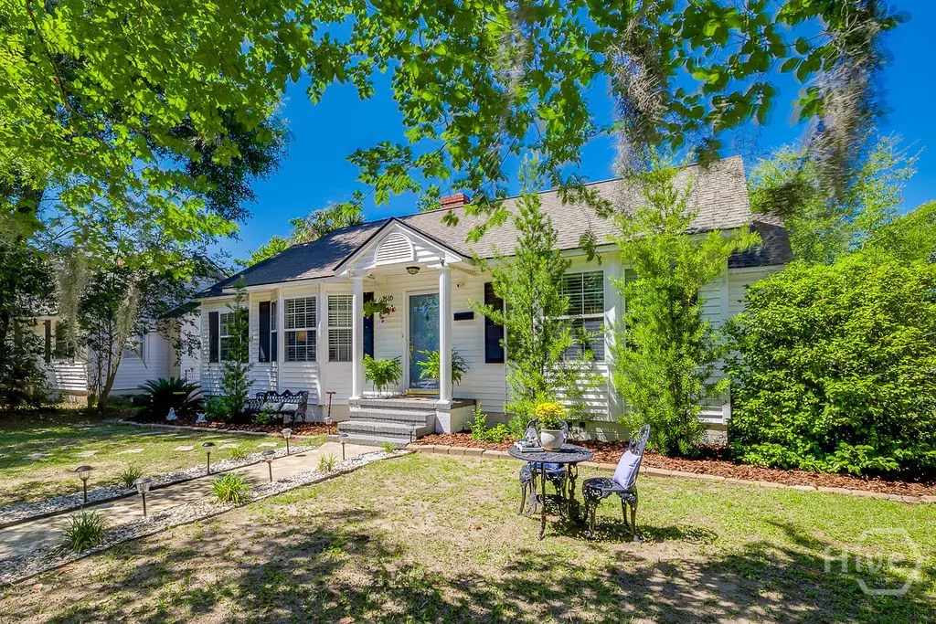 White cottage with a blue door, front porch, and a small bistro set on a sunny lawn under lush green trees.