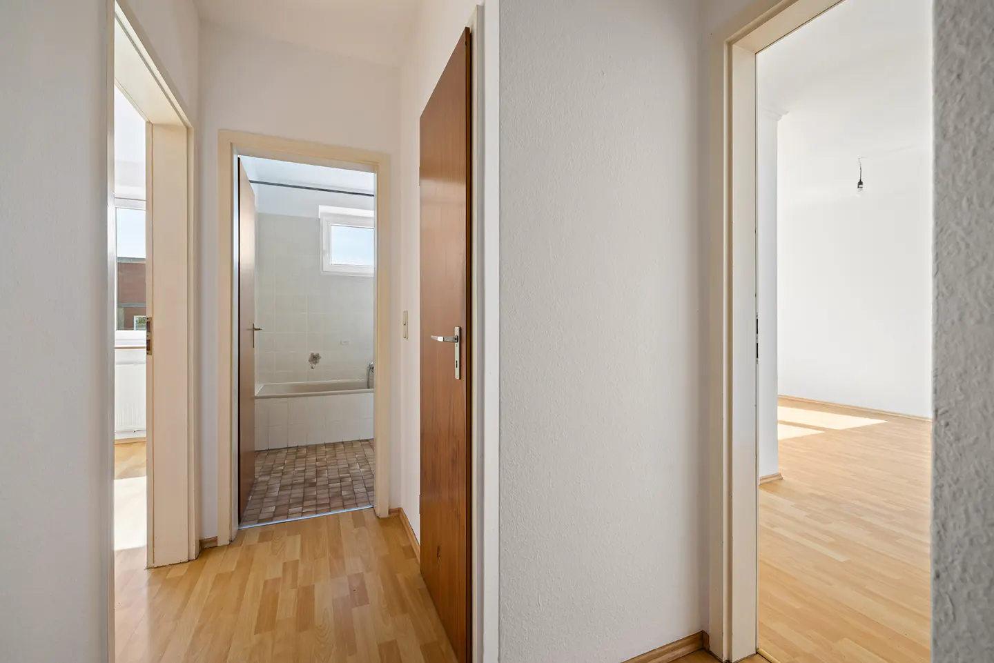 Hallway with wood floors and white walls. Open doors lead to a bathroom and a room with hardwood floors.