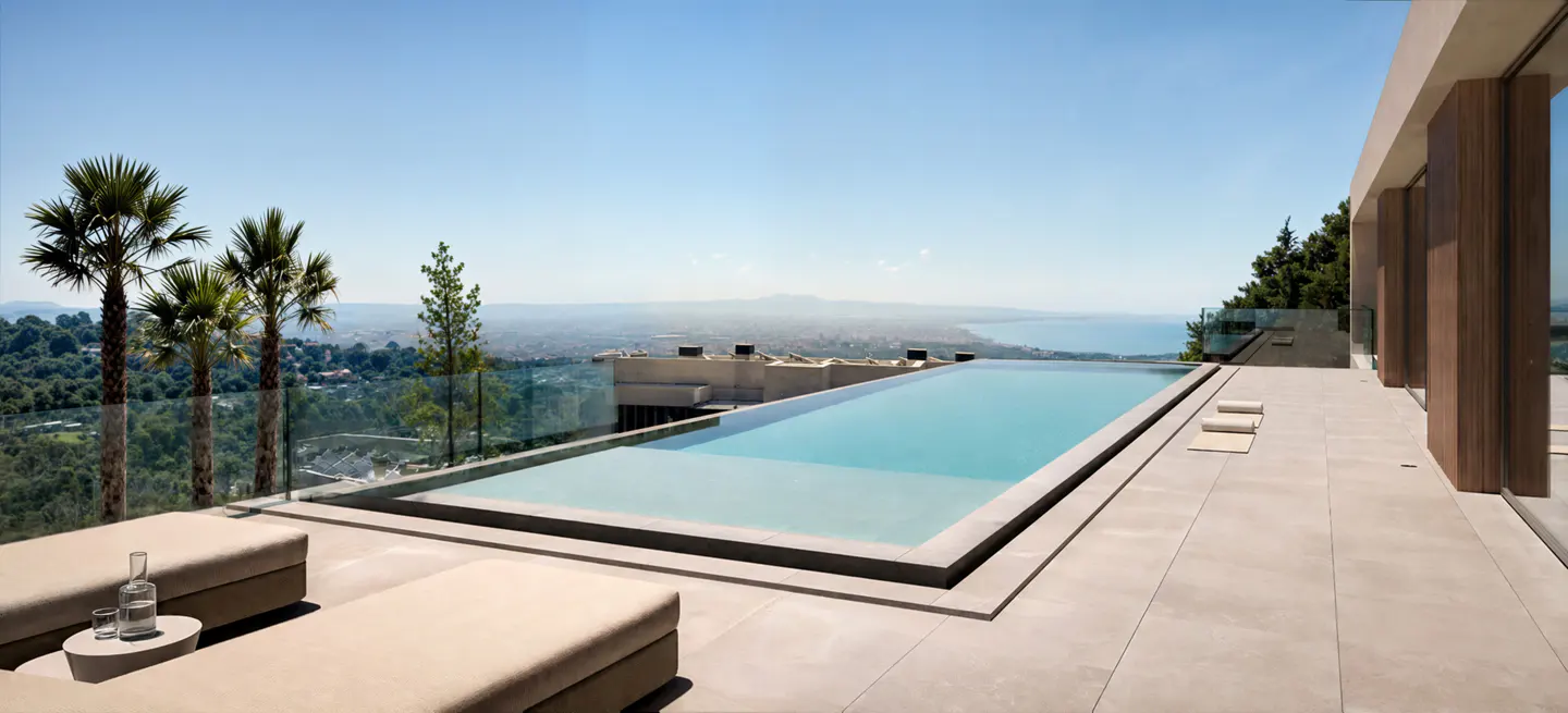 Infinity pool overlooking a scenic view. Lounge chairs, palm trees, and a glass of water are nearby.
