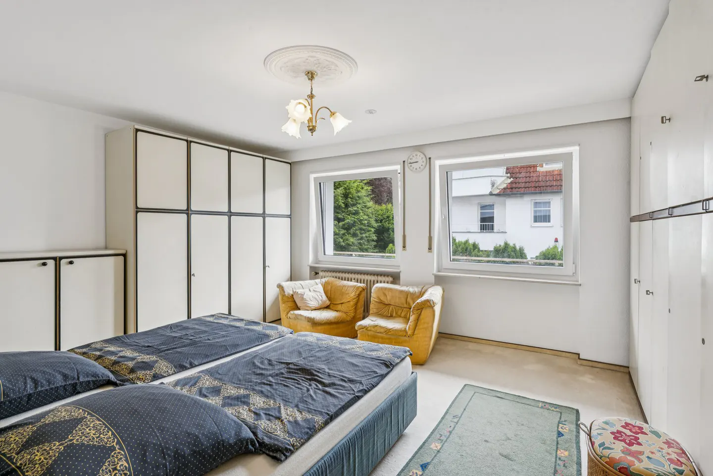 Bedroom with two twin beds, blue patterned bedding, yellow armchairs, and white wardrobes. Windows overlook greenery and a house.