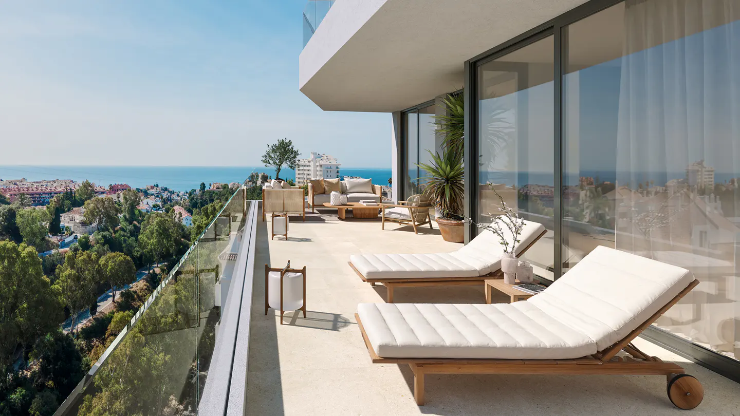Luxury balcony with ocean view. Two white cushioned lounge chairs face the sea. Outdoor seating area with sofa and chairs. Glass railing.