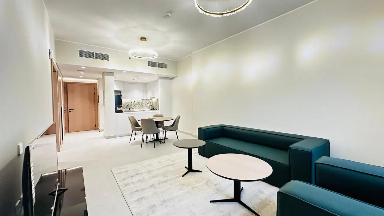 Bright apartment interior with a green sofa, round tables, and a dining area with four chairs. A modern chandelier hangs from the ceiling.