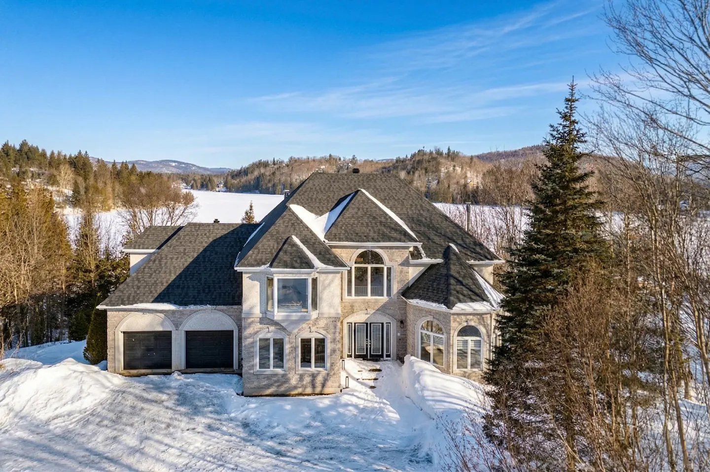 Exterior view of a large, light-stone house with a dark roof and two-car garage in a snowy, wooded landscape.