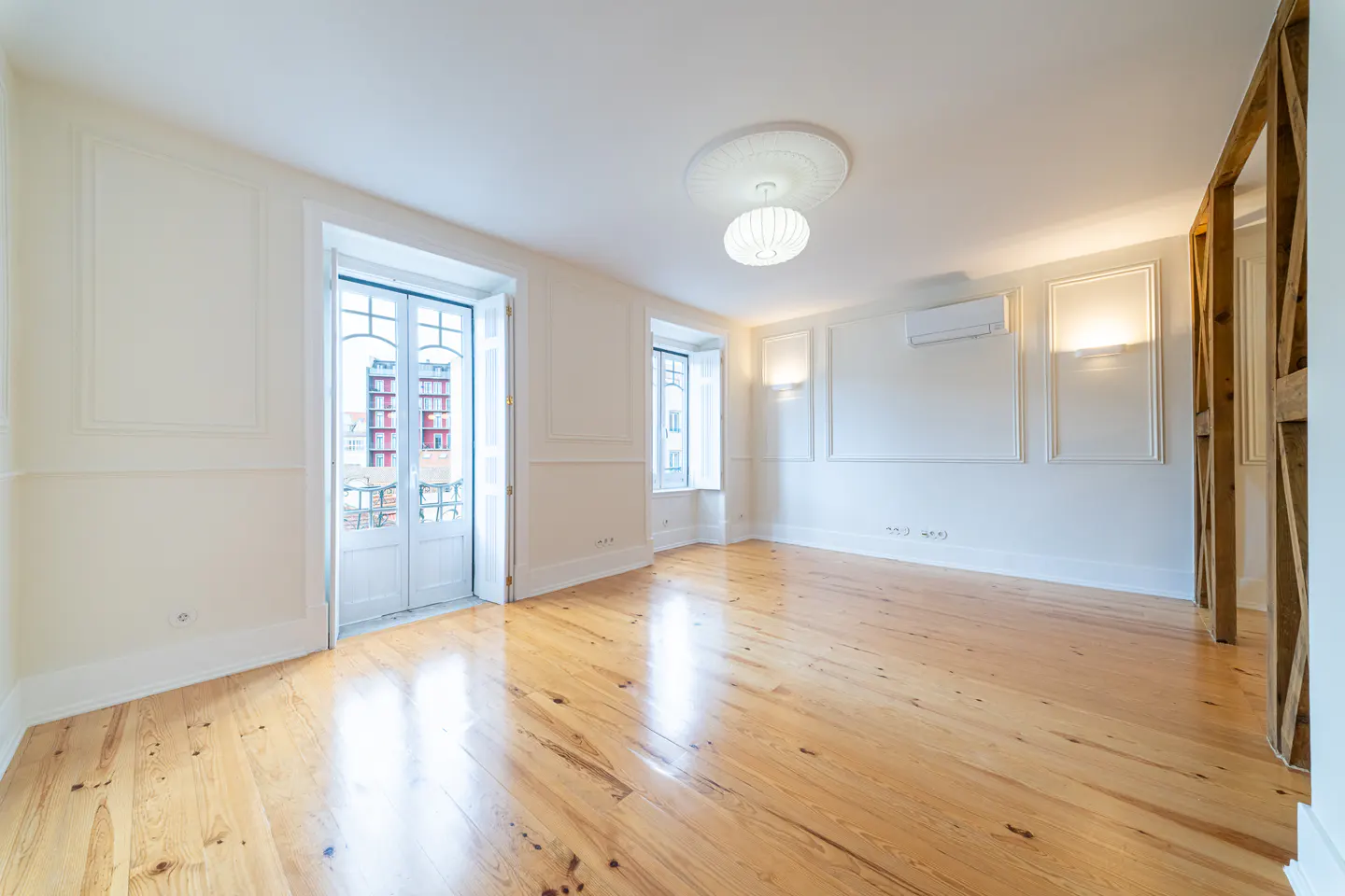 Bright, empty room with light wood floors, white walls, and decorative molding. A white door leads to a balcony. A modern light fixture hangs from the ceiling.