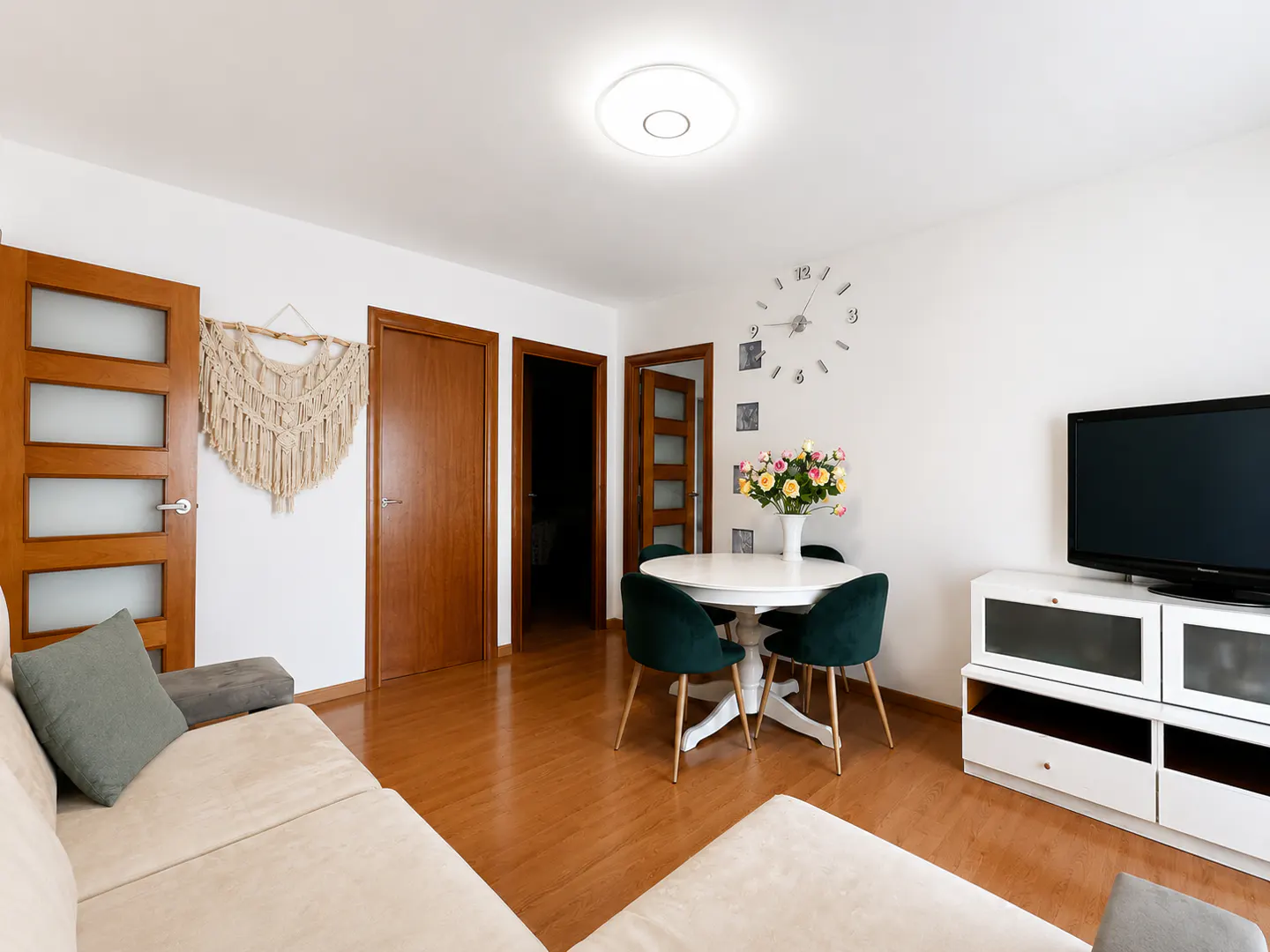 Bright living room with a beige sofa, white walls, wood floors, and a round white table with green chairs. A TV sits on a white cabinet.