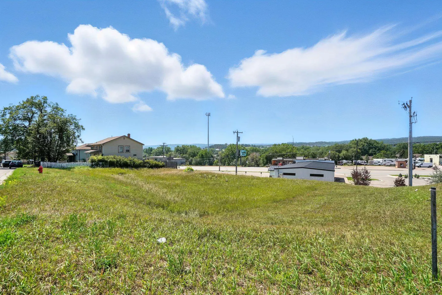 A grassy vacant lot under a blue sky with puffy white clouds. A building and trees are in the background.