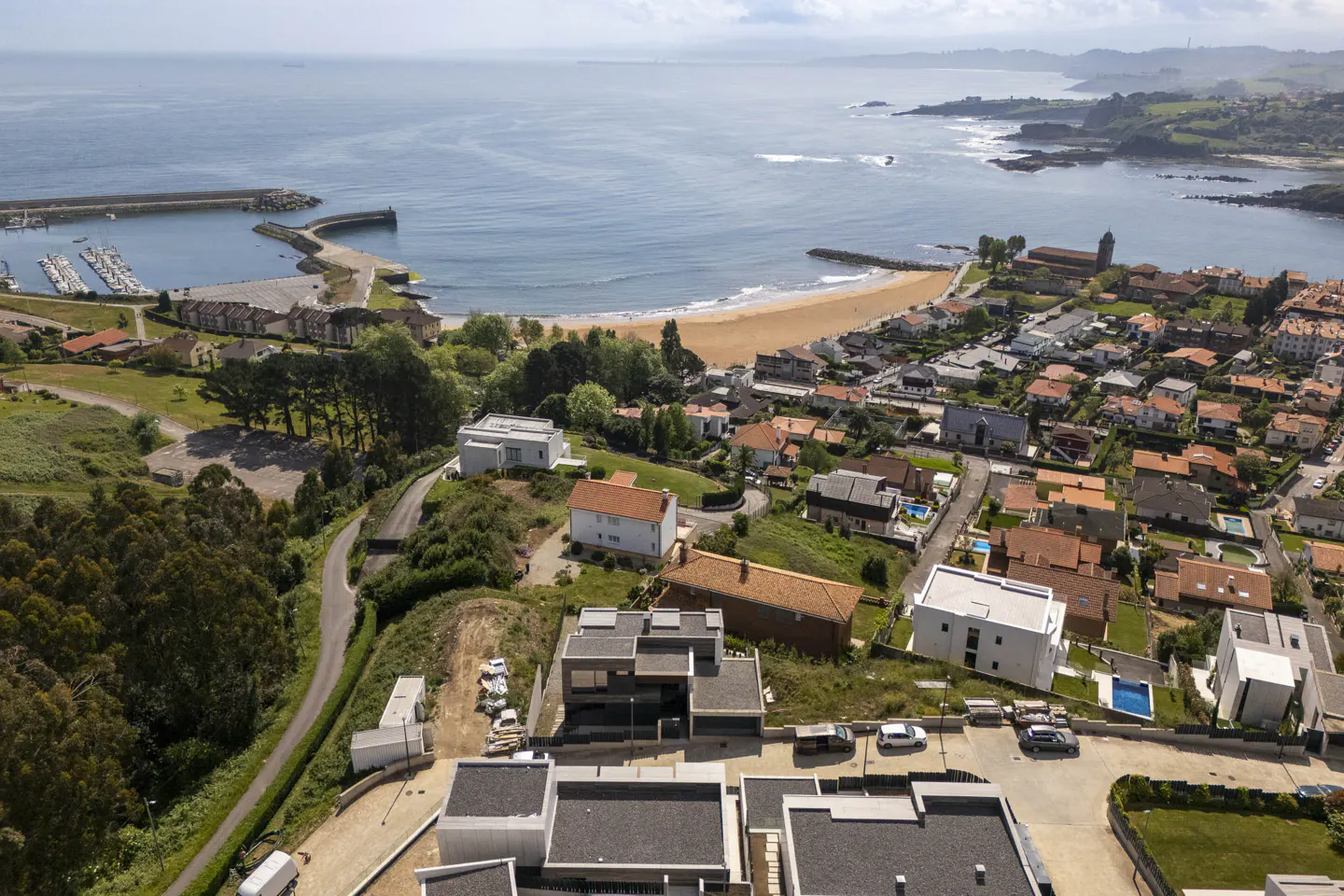 Aerial view of coastal town with modern homes, sandy beach, and blue ocean with a marina in the distance.
