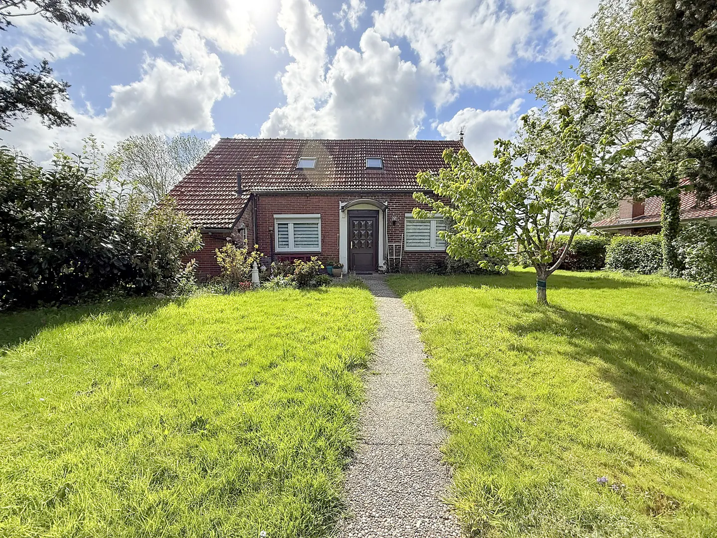 A single-story brick house with a red tile roof and a stone walkway leading to the front door. Green lawn and trees surround the house.