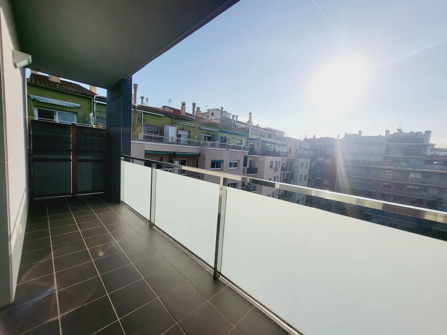 Balcony with dark tile floor and frosted glass railing overlooking city buildings under a bright, sunny sky.