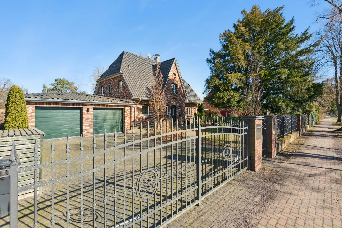 Brick house with a gray roof and green garage doors, seen through a black iron fence on a sunny day.