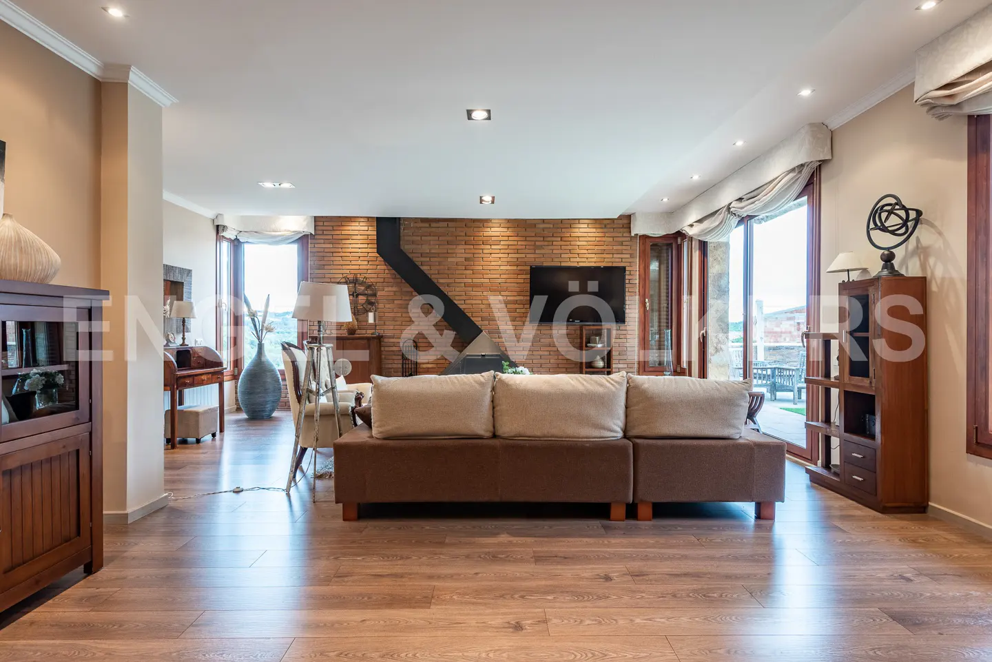 Spacious living room with a brown sectional sofa, brick fireplace wall, flat-screen TV, and wood flooring.