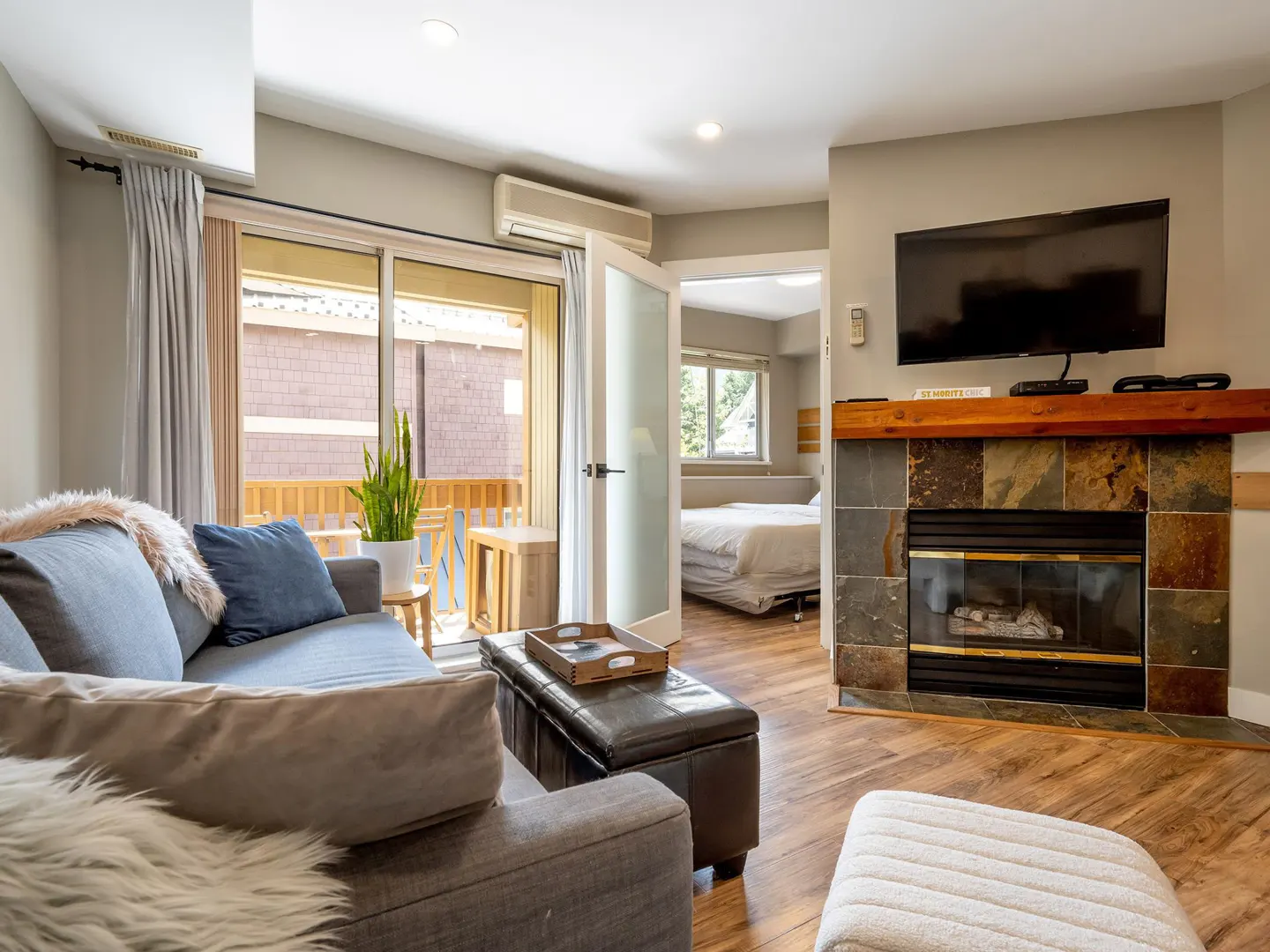 Living room with gray sofa, fireplace, and TV. Balcony with sliding glass doors and bedroom visible through doorway.