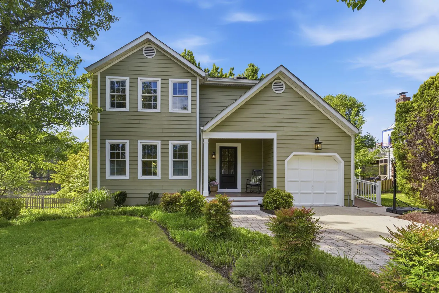 Two-story green house with white trim, a black front door, and an attached garage. A brick walkway leads to the entrance.