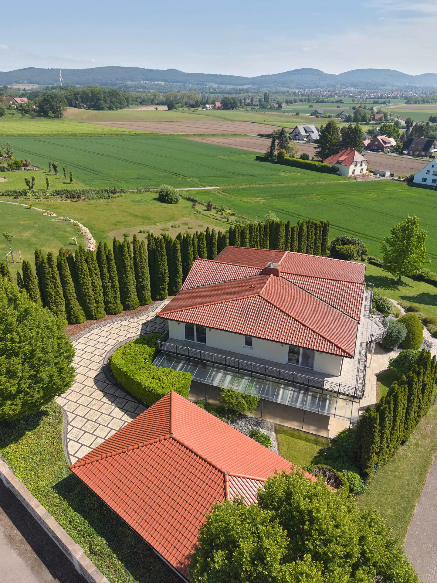 Aerial view of a large, modern house with a red tile roof, surrounded by green lawns, trees, and fields.