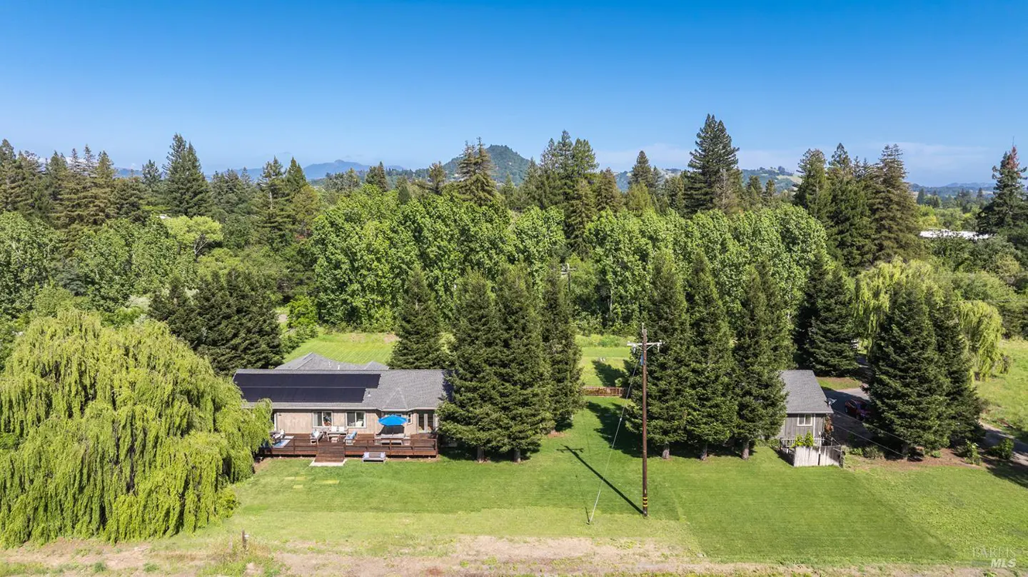 Aerial view of a house with solar panels, a deck, and a blue umbrella, surrounded by green trees and a clear blue sky.