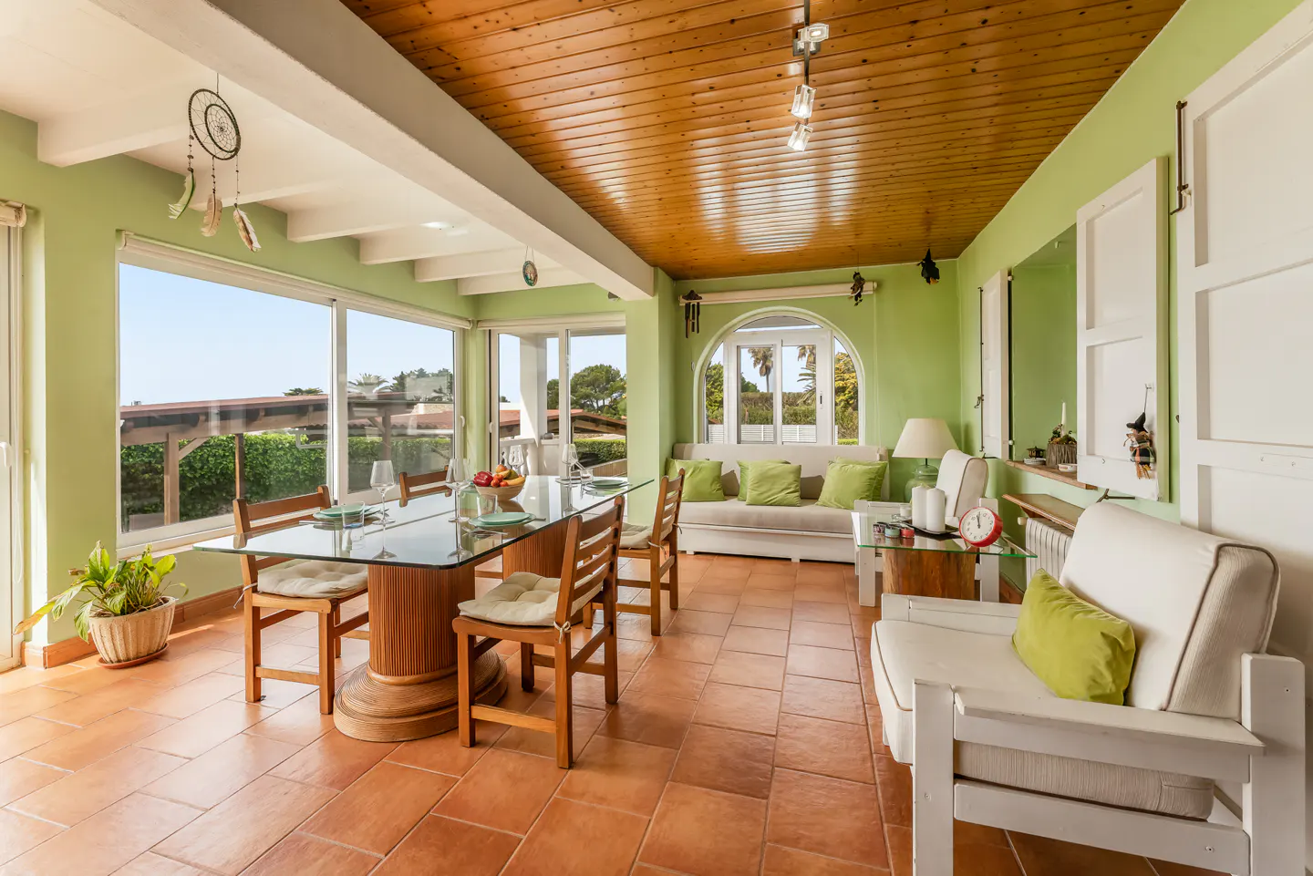 Bright sunroom with green walls, wood ceiling, and terracotta tile floor. A glass-top table is set for a meal. White sofas and chairs offer seating.