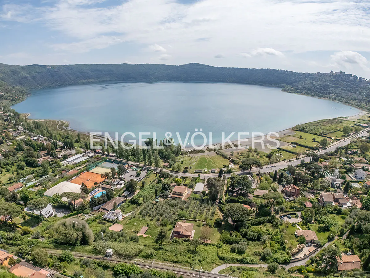 Aerial view of Lake Albano, Italy, surrounded by green hills and residential areas. The water is light blue, and the sky is partly cloudy.