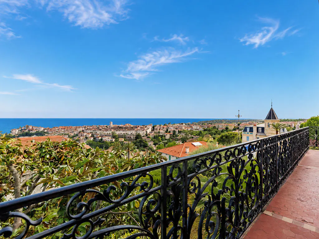 View from a balcony with black ornate railing overlooking a coastal town with red roofs, green trees, and blue sea under a blue sky.