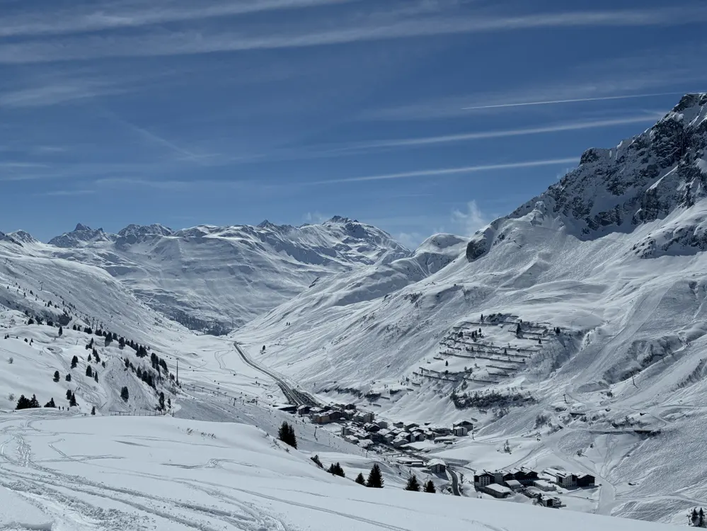 A small village nestled in a vast, snow-covered mountain valley under a bright blue sky with wispy clouds.