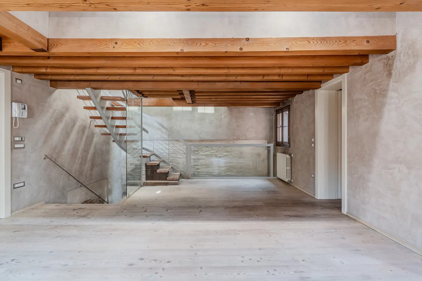 Modern interior with light wood floors, exposed timber ceiling beams, and a floating staircase with glass railings.