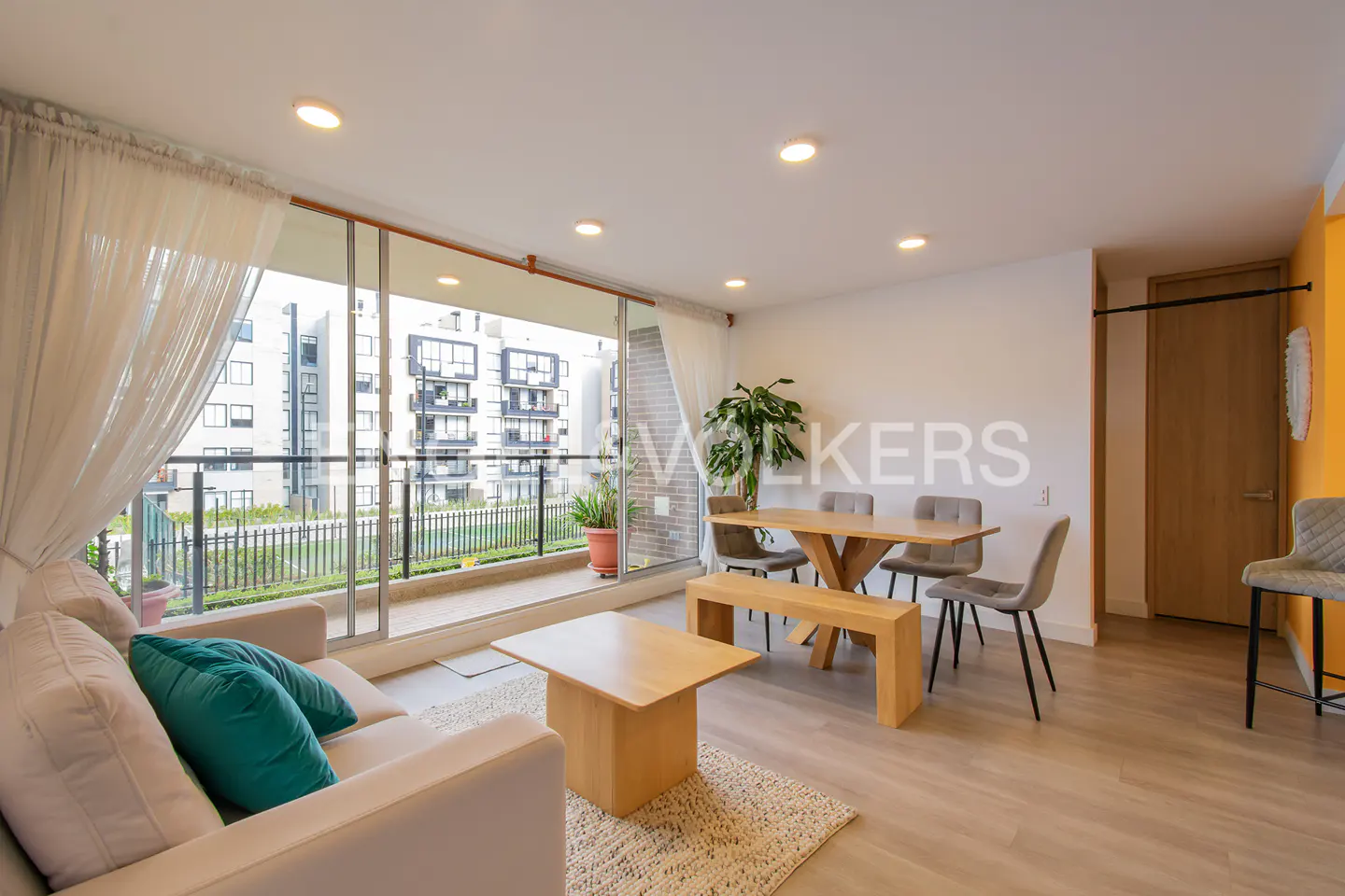 Bright living room with a beige sofa, teal pillows, a wooden table, and a balcony overlooking a building.