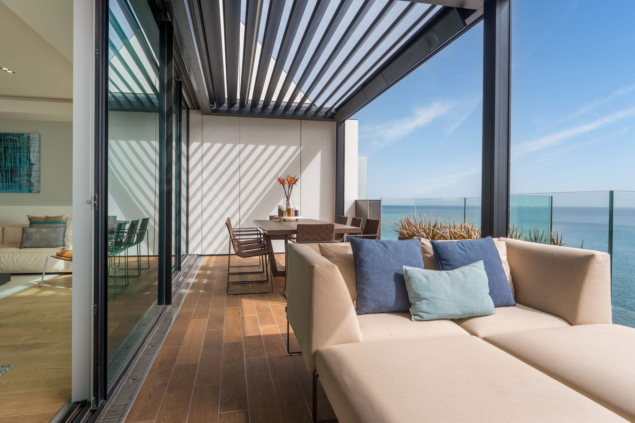 Modern balcony with beige lounge seating, dining table, and louvered roof overlooking the ocean on a sunny day.