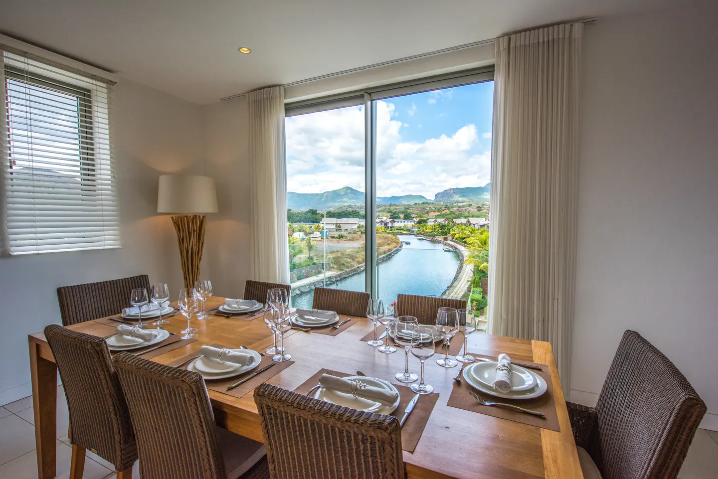 Dining room with a wood table set for eight, wicker chairs, and a large window overlooking a canal and mountains.