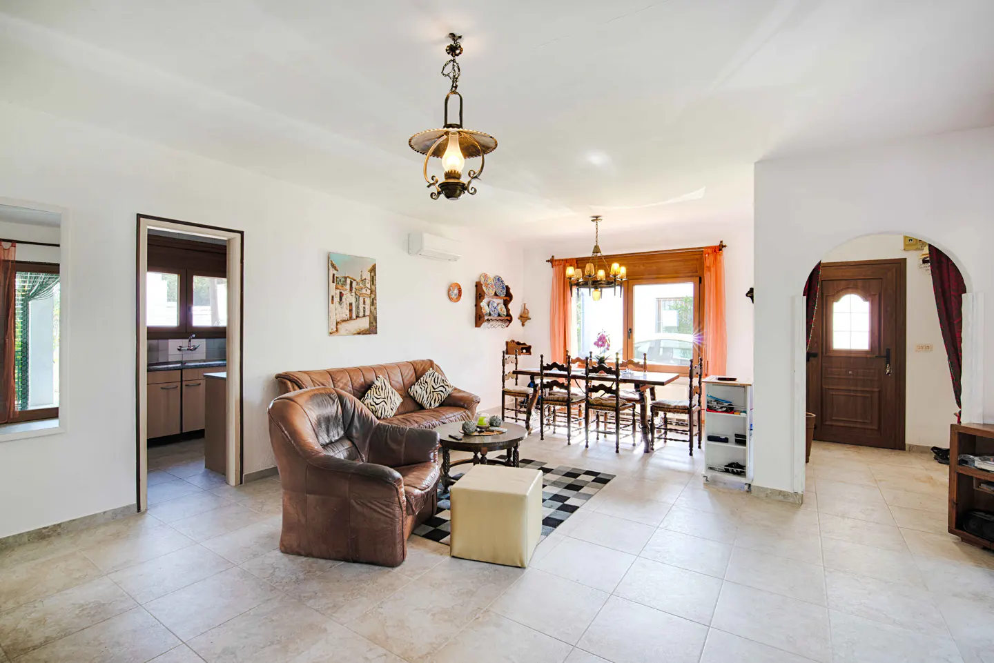 Bright living room with brown leather sofa and chair, round coffee table, and dining table with chairs. Tiled floor and white walls.