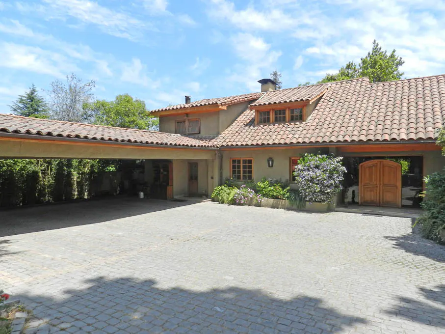 Exterior view of a tan house with a red tile roof, a carport, and a cobblestone driveway.