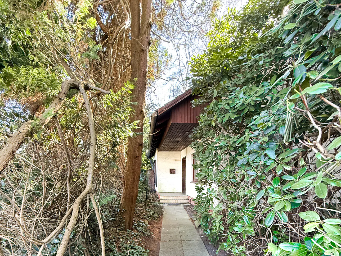Exterior view of a white house with a brown roof, partially obscured by trees and bushes. A stone path leads to the entrance.