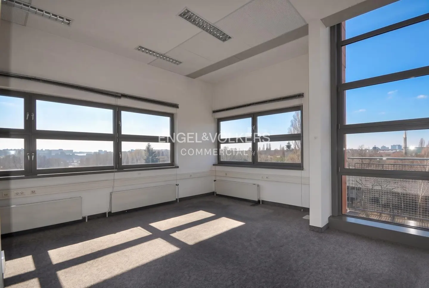 Bright, empty office space with large windows showing a city view. Gray carpet and white walls. Engel & Volkers logo visible.