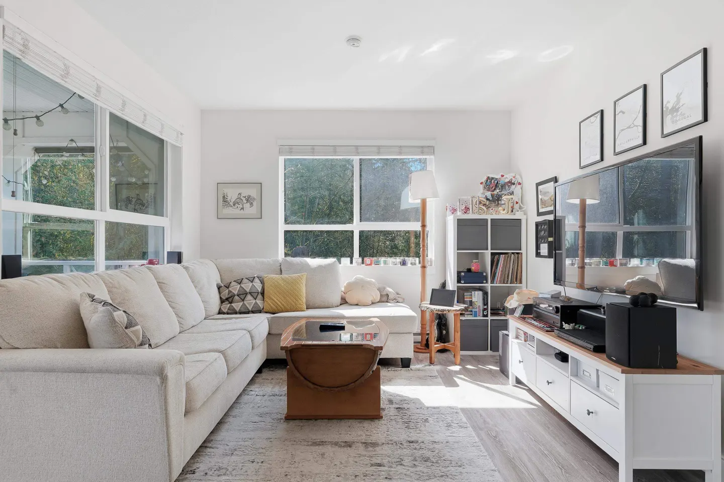Bright living room with a white sectional sofa, wood coffee table, and large windows overlooking greenery. A TV and shelving unit are against the wall.