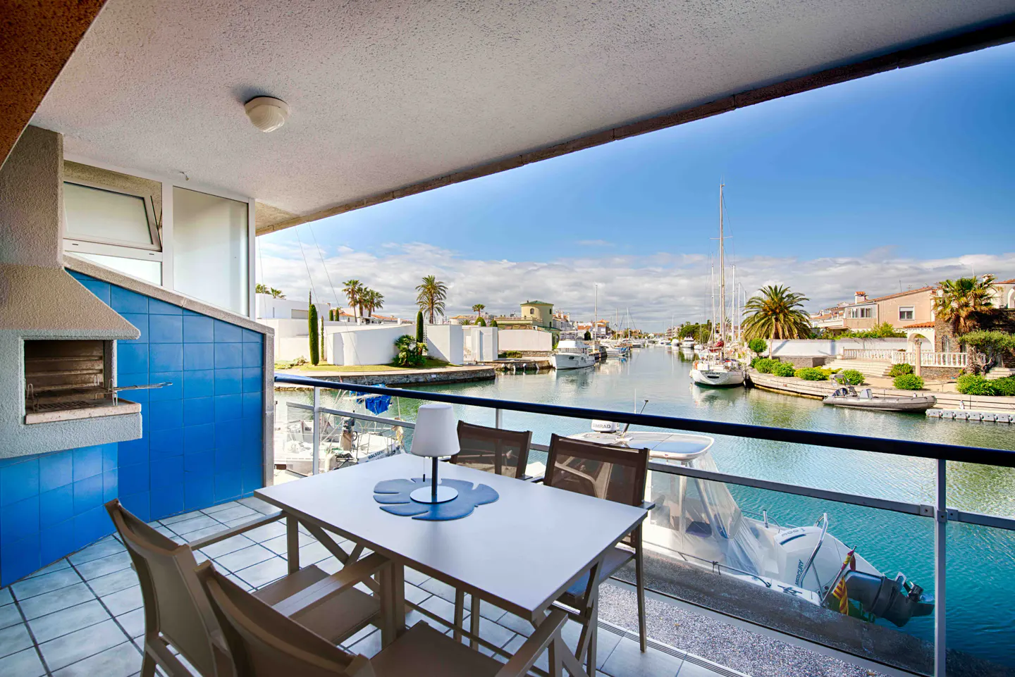Balcony with table, chairs, and grill overlooking a canal with boats and waterfront homes on a sunny day.