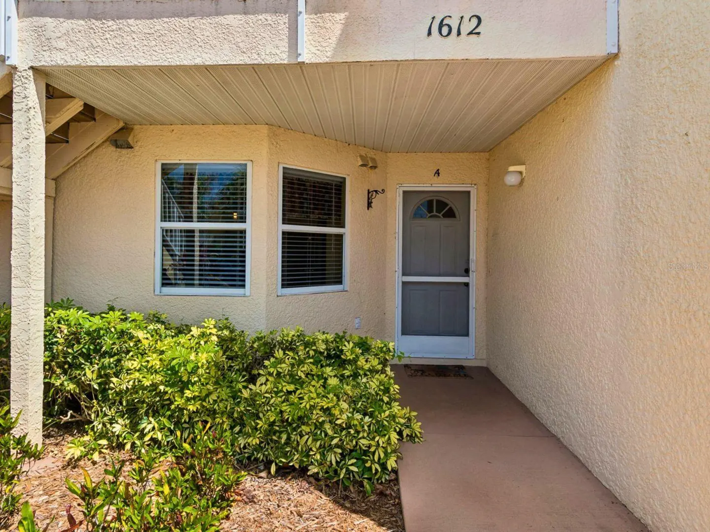 Exterior view of a tan stucco condo with the address 1612 above the door. Windows with blinds are to the left of the door, and green bushes are in front.