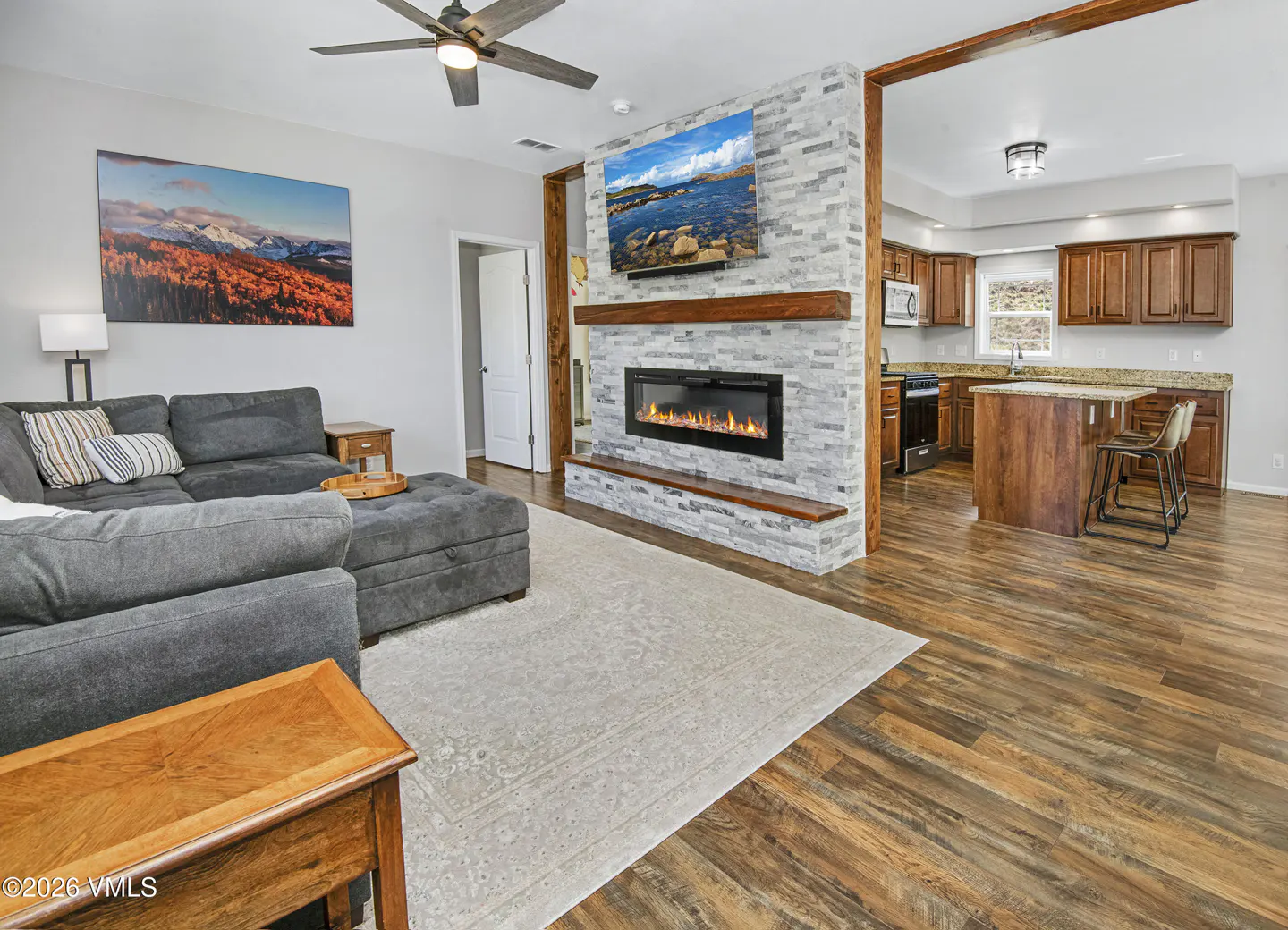Living room with gray sectional, stone fireplace, and TV. Open kitchen with wood cabinets and island. Hardwood floors throughout.