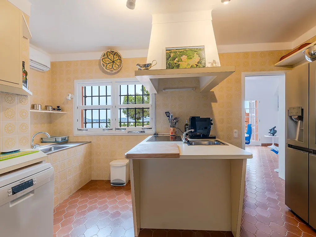 A bright kitchen with yellow patterned walls, terracotta floors, and white cabinets. A window overlooks the sea.