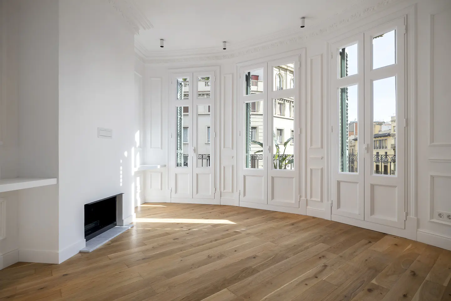 Bright, empty room with white walls, hardwood floors, a modern fireplace, and large windows overlooking a city street.