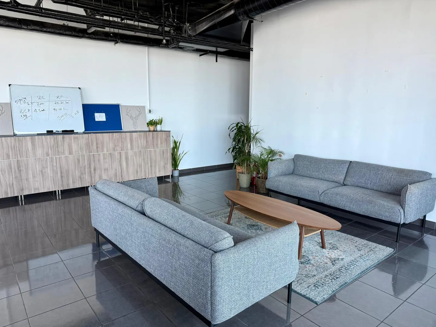 Bright office space with two gray sofas, a wood coffee table on a rug, and plants. A whiteboard is on the left.