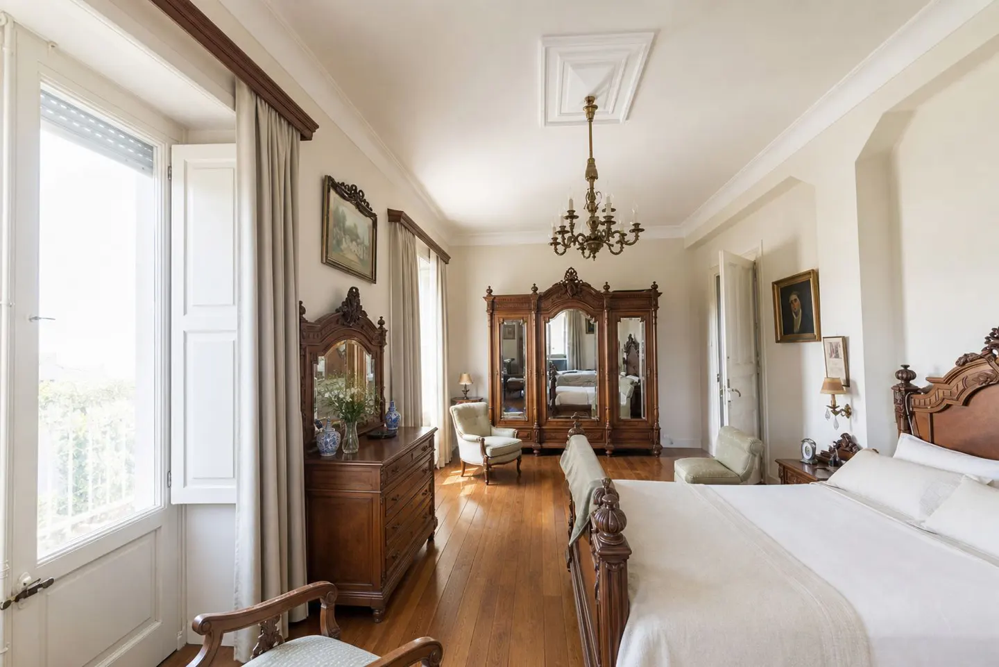 Bright bedroom with hardwood floors, white walls, and antique wood furniture including a bed, dresser, and wardrobe. A chandelier hangs from the ceiling.