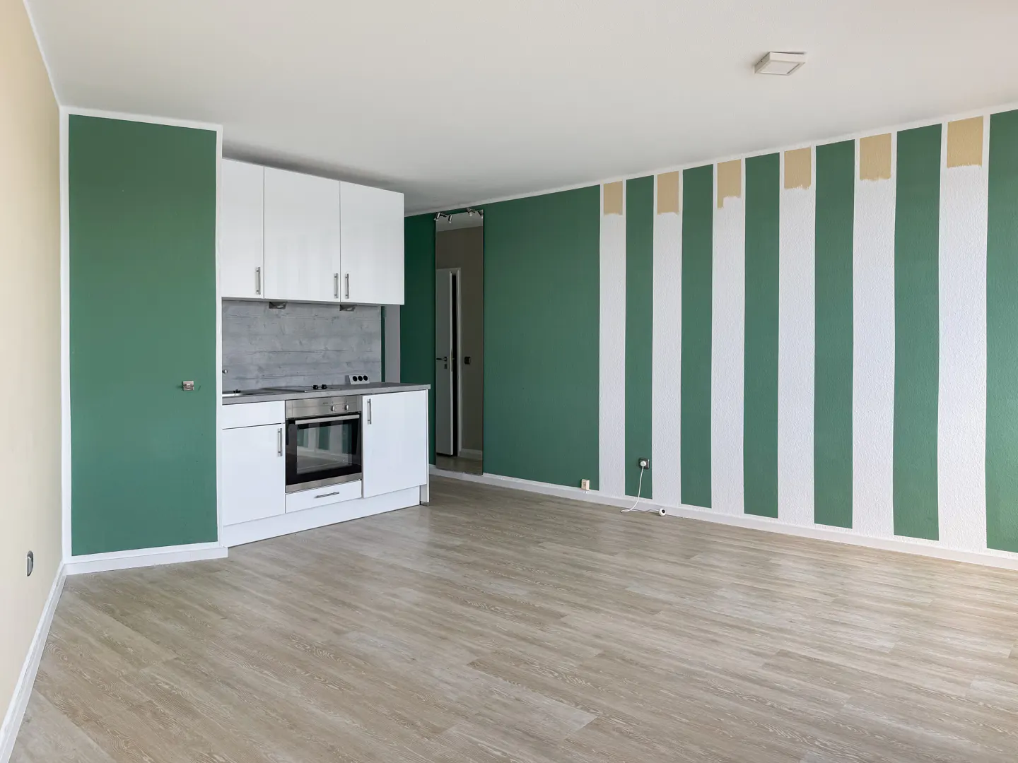 Open-concept apartment with green and white striped walls, light wood floors, and a small white kitchen.