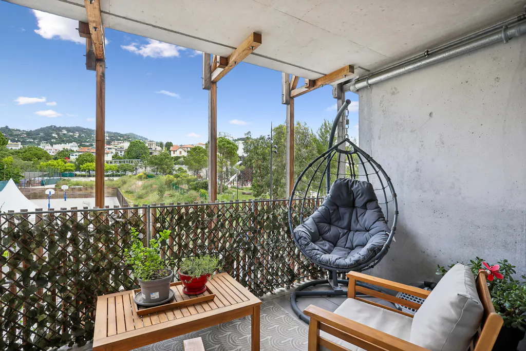 Balcony view with a hanging chair, wooden table with plants, and a wooden chair. The background shows a landscape with trees and buildings.