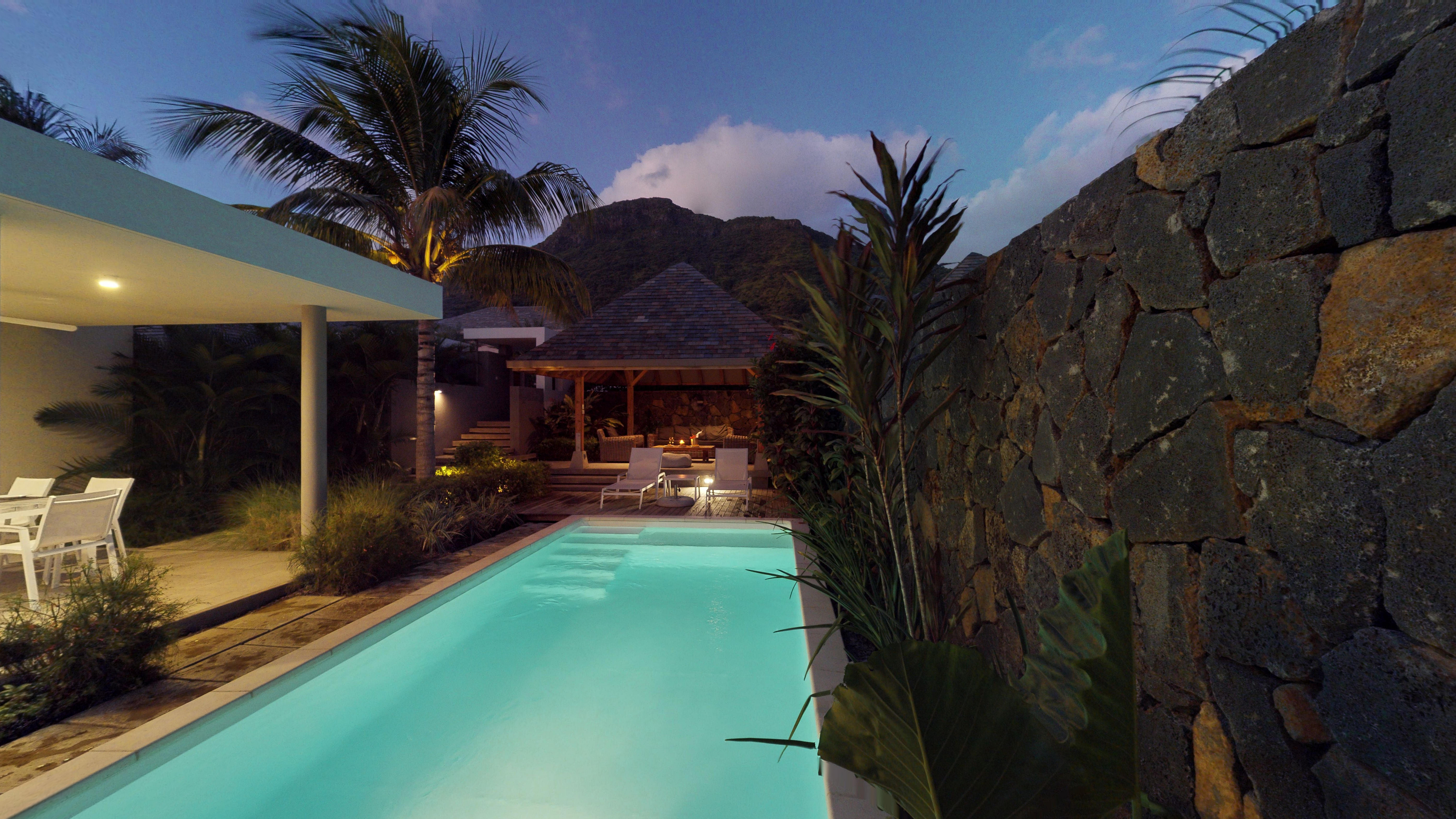 Outdoor pool at dusk with a gazebo, lounge chairs, and a stone wall. Palm trees and a mountain are in the background.