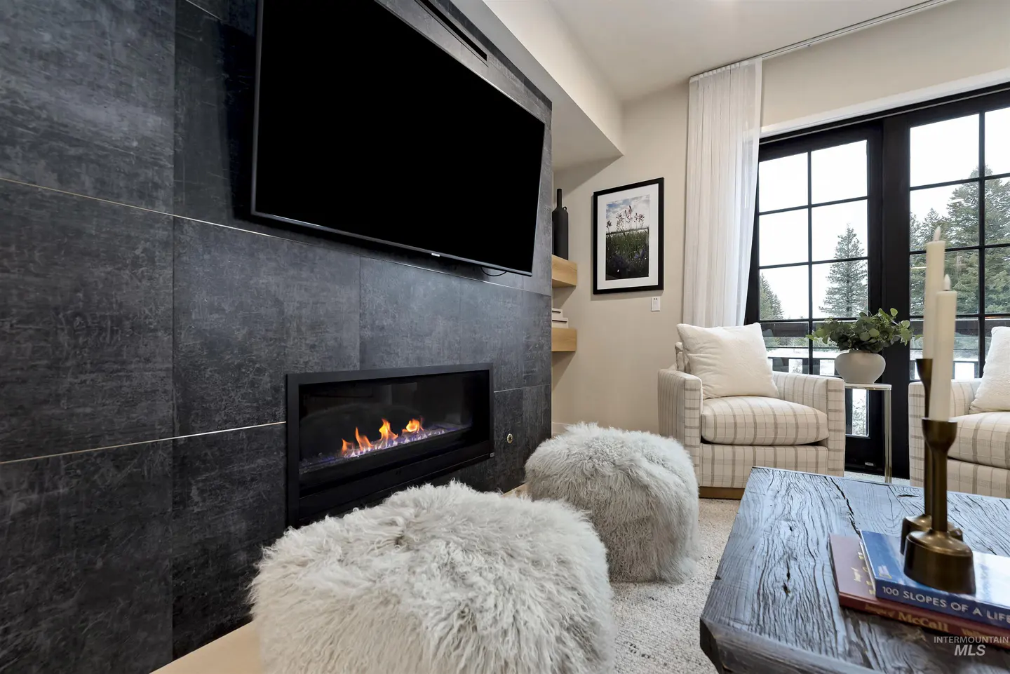 Living room with a black tiled fireplace, TV, and a modern gas fire. Two white furry ottomans sit in front of the fireplace. Plaid armchairs face a rustic coffee table.