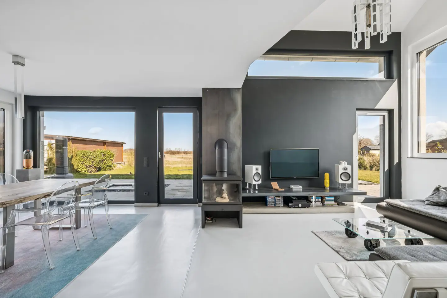 Bright, modern living room with white floors, gray walls, and large windows. A dining table with clear chairs sits near a door to the outside.