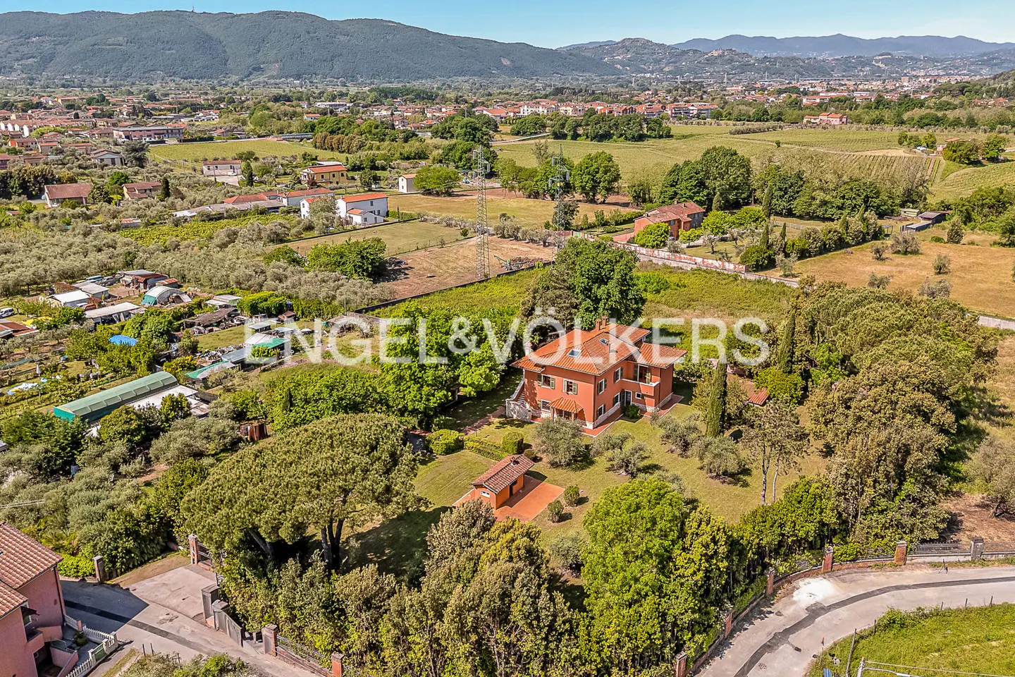 Aerial view of a two-story orange house with a red tile roof, surrounded by green trees and fields, under a blue sky.