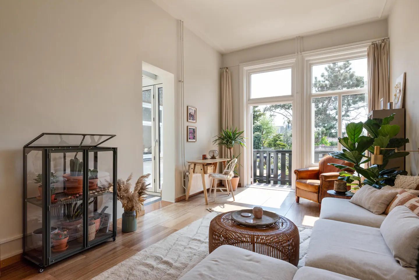 Bright living room with a white sofa, brown armchair, and a glass terrarium filled with plants. A desk sits near a balcony with a view of trees.