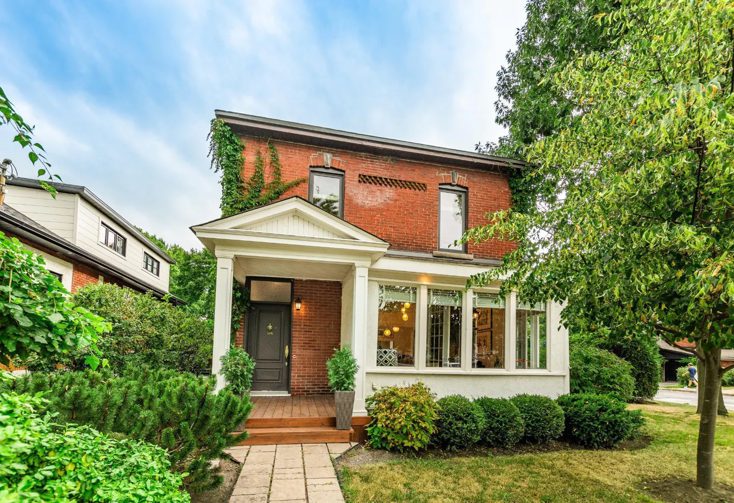 Two-story red brick house with a white porch, green bushes, and trees under a blue sky.