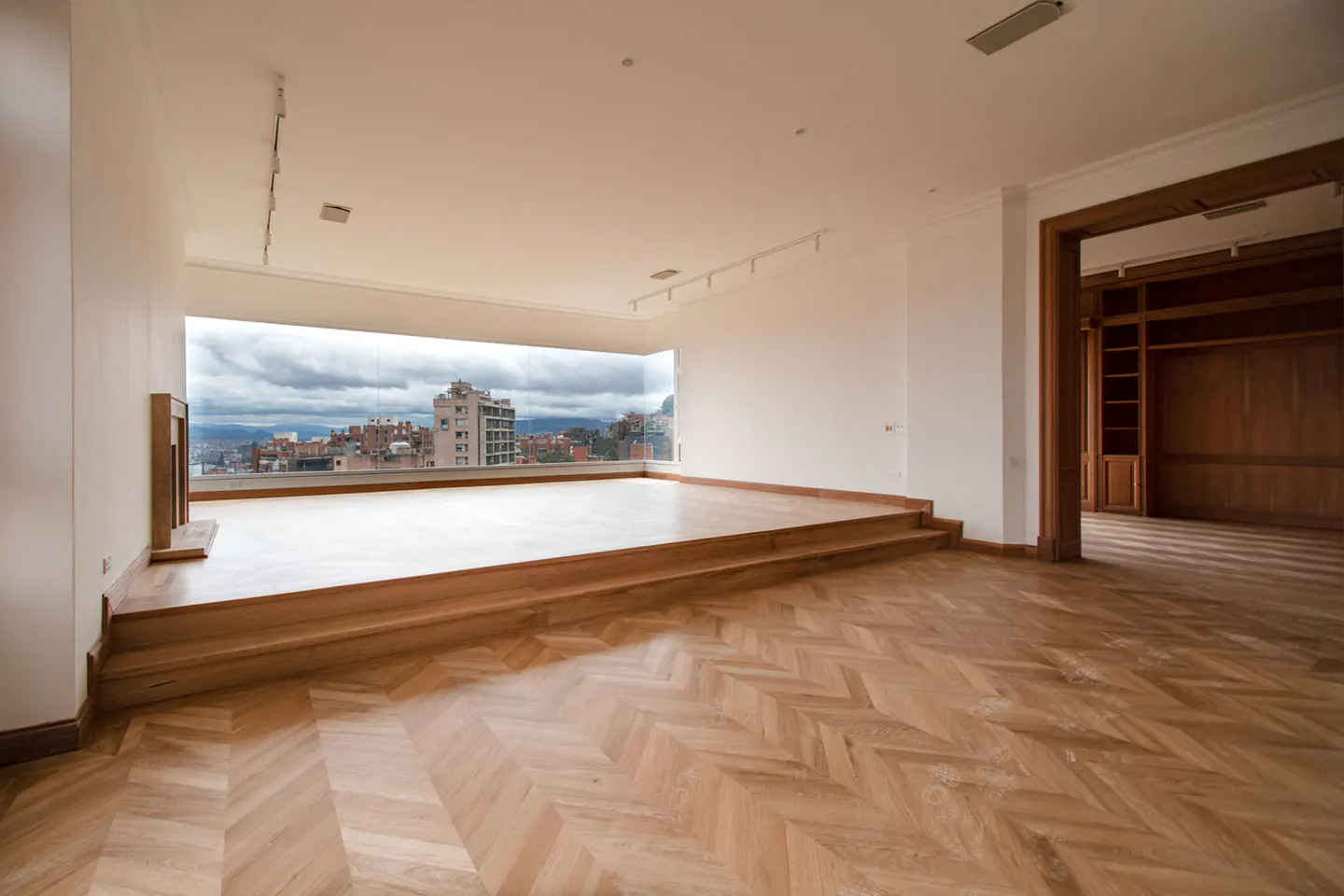 Bright, empty room with herringbone wood floors and a large window overlooking a city skyline. A wood-paneled doorway leads to another room.