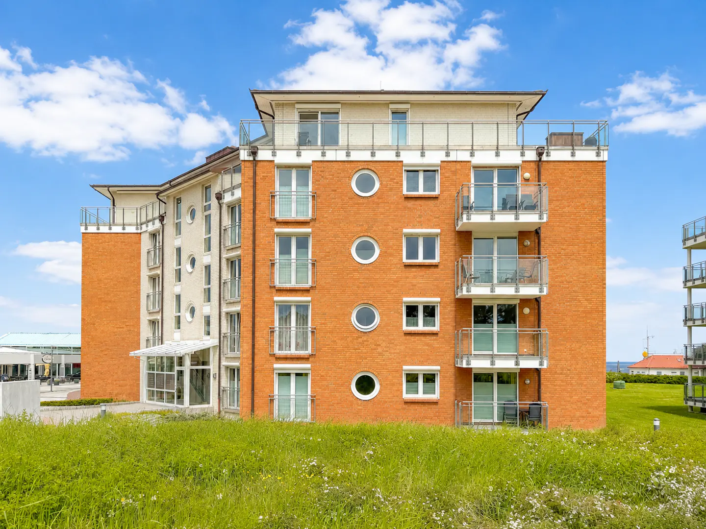 Modern orange brick apartment building with balconies and circular windows, set against a blue sky with green grass.