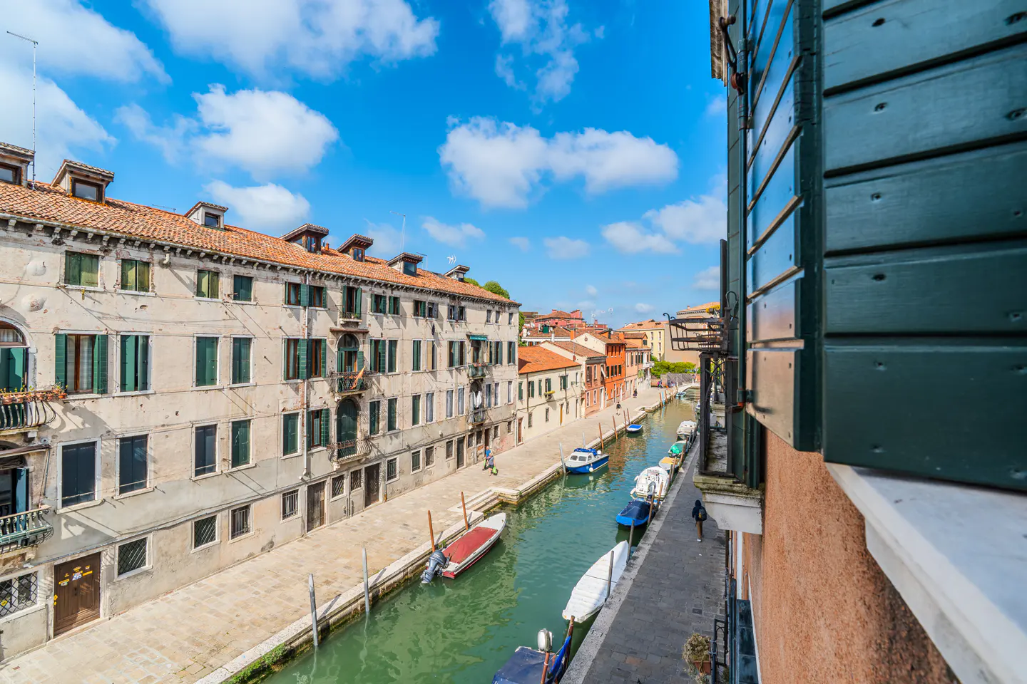 View of a Venice canal with boats, buildings with green shutters, and a blue sky with white clouds.